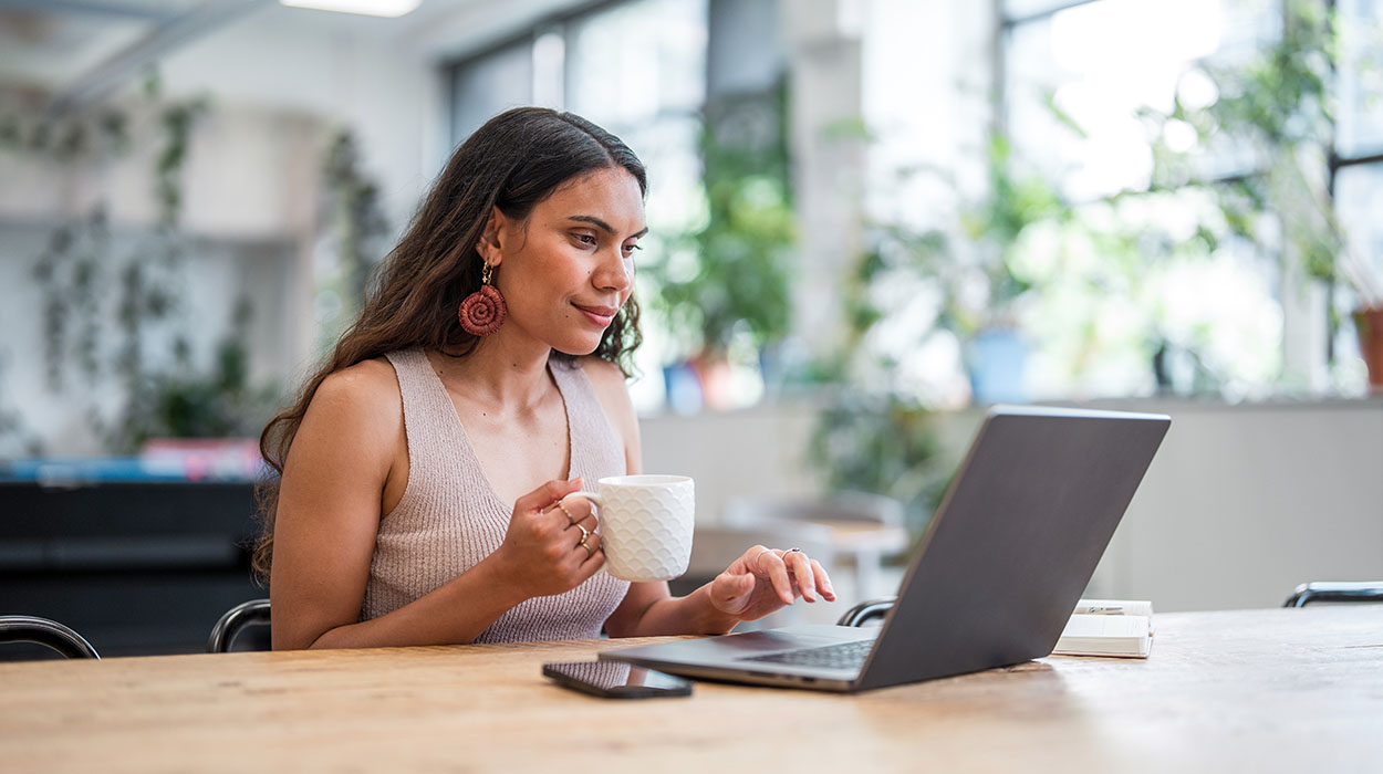 A woman with long dark hair sits at a wooden table, holding a white mug and looking at her laptop. She is indoors, surrounded by plants, with a phone and a notebook on the table.